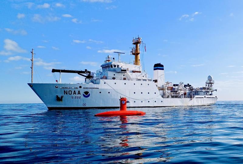 A white NOAA ship with an orange uncrewed surface vehicle on the water in the foreground