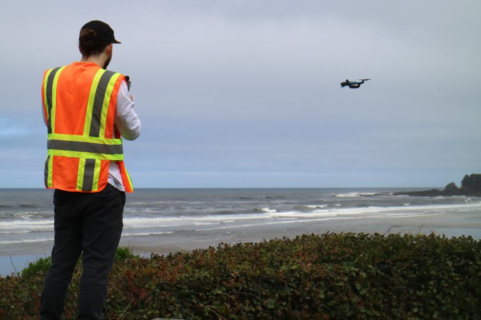 A man in an orange and yellow safety vest stands on a beach with his back to the camera. He is piloting a small drone that is flying in the distance over the sand.