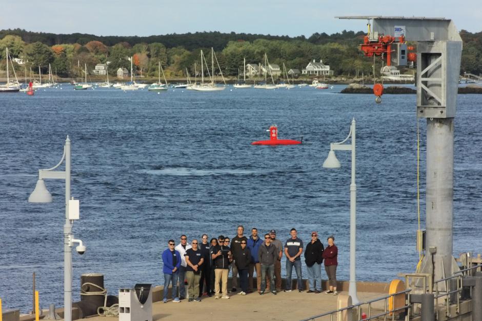 The Uncrewed Systems Operations Center team poses for a photo taken via uncrewed aircraft as the DriX uncrewed surface vehicle transits behind them.