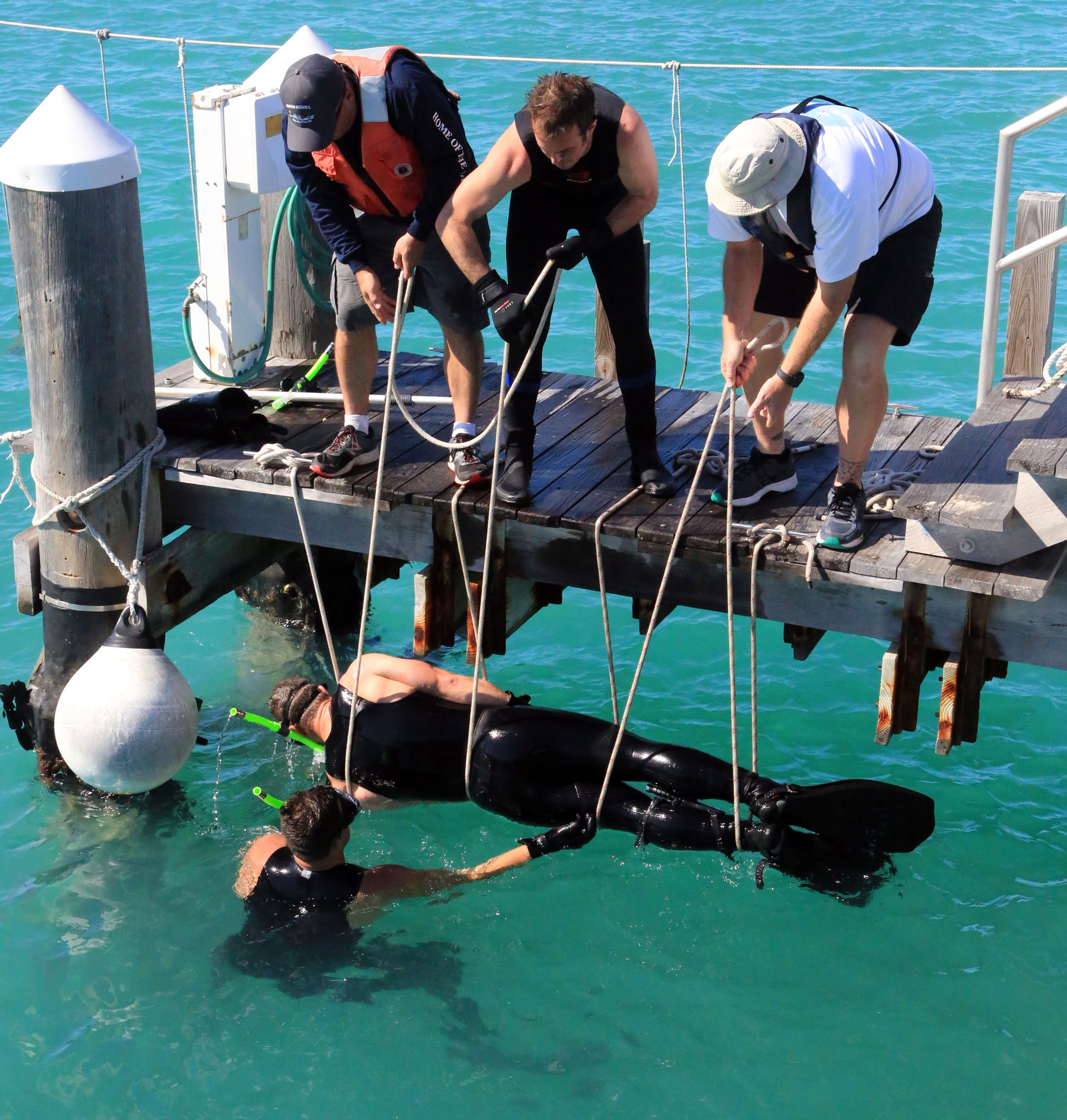three students standing on a pier haul a diver from the water using a rope