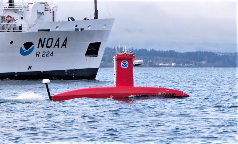 A bright orange submarine-shaped vehicle on the water with a NOAA ship behind it