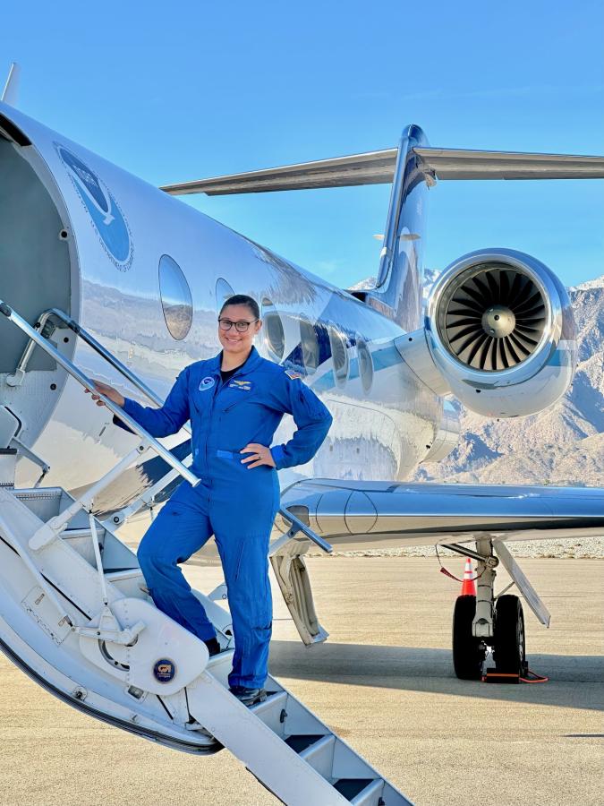 A NOAA Corps officer in a blue flightsuit standing on the stairs to a NOAA jet