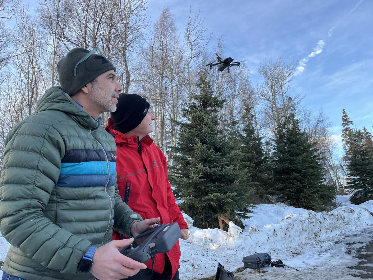 Two uncrewed aircraft operators dressed in winter clothes observe an uncrewed aircraft flying over a snowy patch of trees.