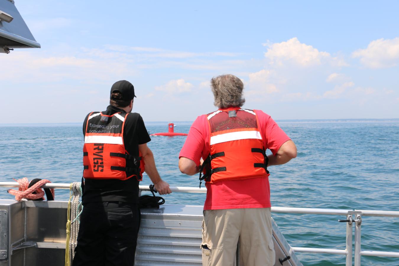 Two people in orange life jackets with their backs to the camera stare out at the ocean as a red uncrewed surface vehicle drives by.