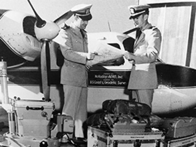 Two male uniformed officers standing next to a twin-engine airplane in 1956