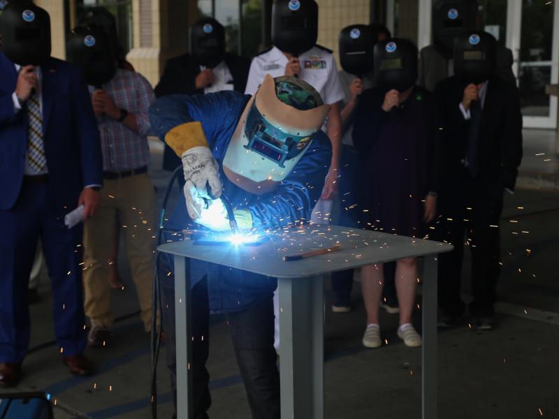 A welder working on a metal plate while a group of people look on