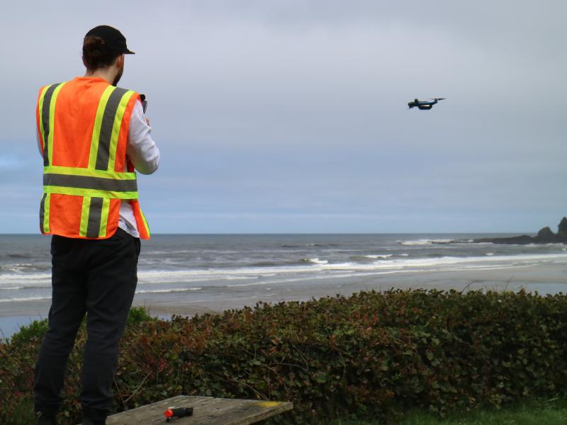 A man in an orange and yellow safety vest stands on a beach with his back to the camera. He is piloting a small drone that is flying in the distance over the sand.