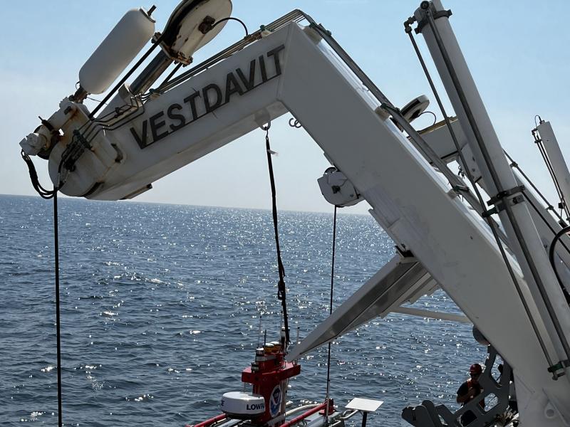 A gray davit arm extends over the edge of a ship. The DriX deployment gondola with a red DriX are being lowered by the davit.