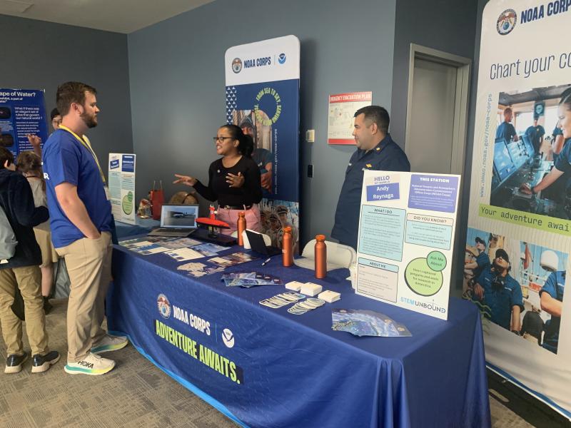 OMAO personnel stand behind a blue table with resources on it. They engage with event attendees who pass by.