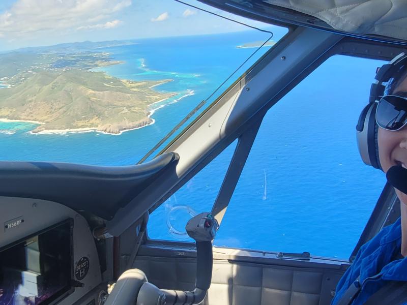 NOAA Corps pilot smiles in flight station of NOAA Twin Otter with view of St. Croix water and landscape in the background
