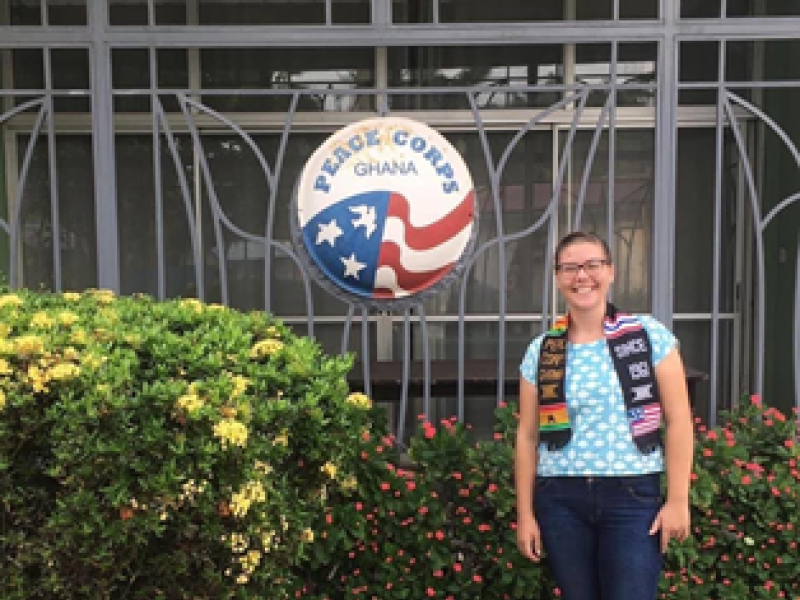 A woman stands in front of a Peace Corps logo, smiling with a Peace Corps stole on.
