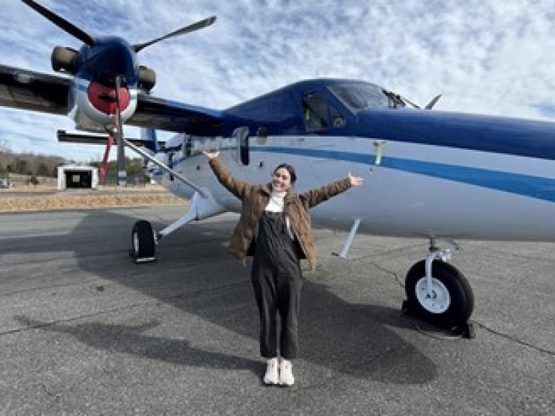 An OMAO Knauss Fellow stands in front of a NOAA Twin Otter plane, posing with a smile and outstretched arms.