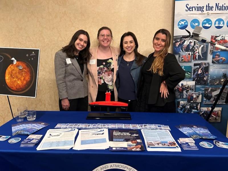 Four women in business casual attire stand in front of a table with NOAA outreach materials on it.
