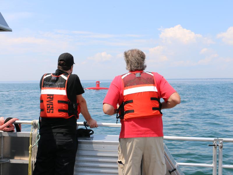 Two people in orange life jackets with their backs to the camera stare out at the ocean as a red uncrewed surface vehicle drives by.
