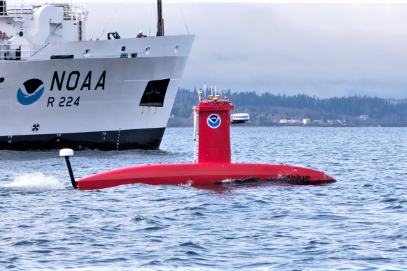 A bright orange submarine-shaped vehicle on the water with a NOAA ship behind it
