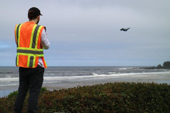 A man in an orange and yellow safety vest stands on a beach with his back to the camera. He is piloting a small drone that is flying in the distance over the sand.