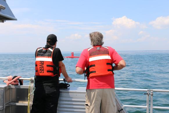 Two people in orange life jackets with their backs to the camera stare out at the ocean as a red uncrewed surface vehicle drives by.