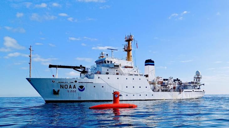 A white NOAA ship with an orange uncrewed surface vehicle on the water in the foreground