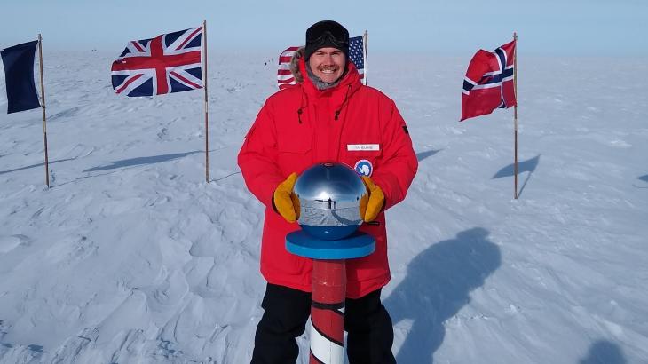 A NOAA Corps officer in a bright orange jacket at the striped South Pole marker