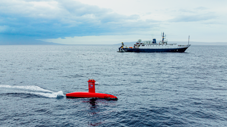 A red uncrewed marine system transits on top of the ocean with the E/V Nautilus in the background.