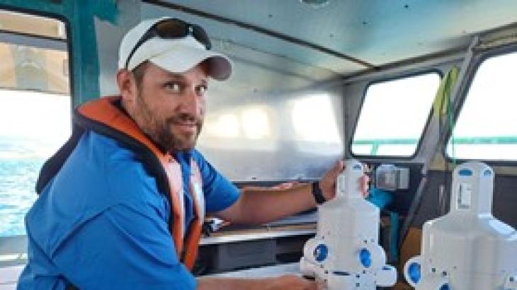A NOAA scientist poses for the camera in a blue sun shirt, white hat, and orange life jacket while in the cabin of a boat. They are holding a small, white uncrewed marine systems called the Hydrus.