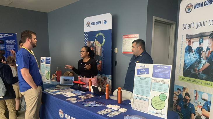 OMAO personnel stand behind a blue table with resources on it. They engage with event attendees who pass by.