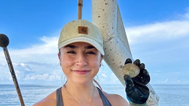 A woman smiles at the camera on a boat while holding a clam.