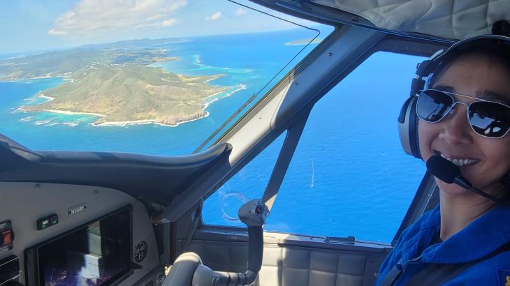 NOAA Corps pilot smiles in flight station of NOAA Twin Otter with view of St. Croix water and landscape in the background