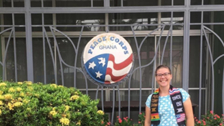 A woman stands in front of a Peace Corps logo, smiling with a Peace Corps stole on.