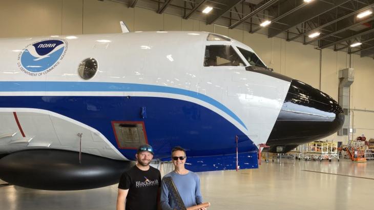 Two individuals stand in front of a NOAA Hurricane Hunter plane with a uncrewed aircraft in their hands.