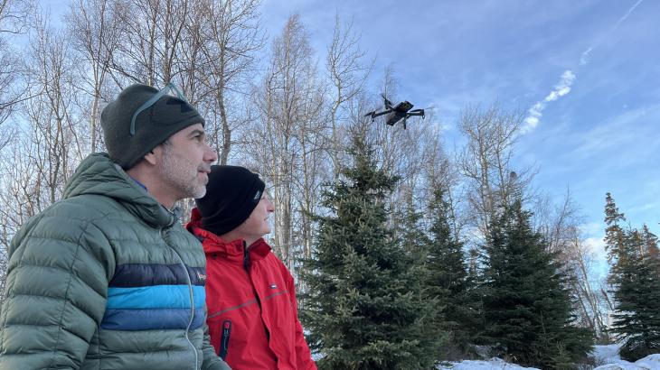 Two uncrewed aircraft operators dressed in winter clothes observe an uncrewed aircraft flying over a snowy patch of trees.