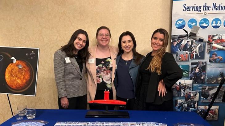 Four women in business casual attire stand in front of a table with NOAA outreach materials on it.