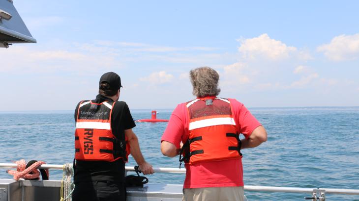 Two people in orange life jackets with their backs to the camera stare out at the ocean as a red uncrewed surface vehicle drives by.