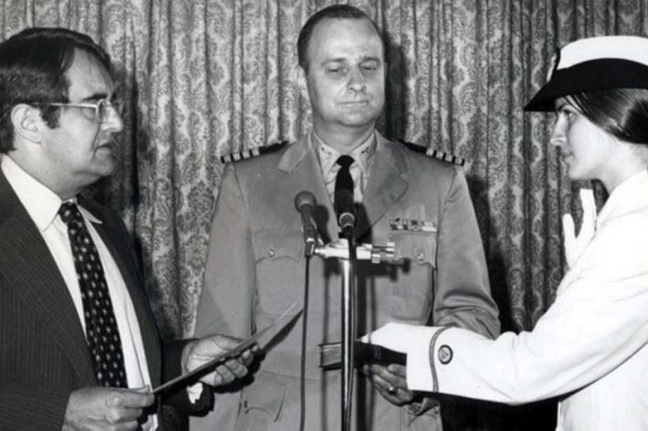 A black-and-white photograph of Ensign Pamela Chelgren was sworn in by Secretary of Commerce Peter G. Peterson on July 6, 1972. Her father, Navy Captain John Chelgren is in the center holding the Bible