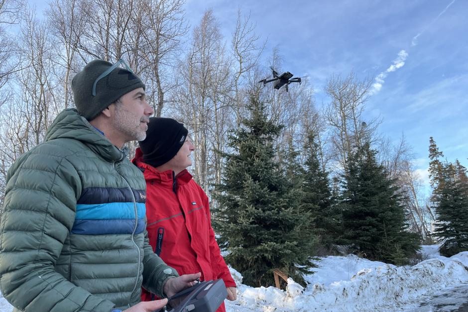 Two uncrewed aircraft operators dressed in winter clothes observe an uncrewed aircraft flying over a snowy patch of trees.