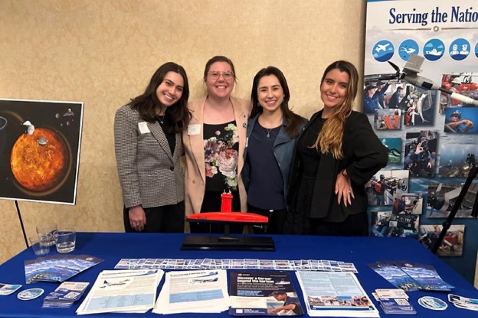 Four women in business casual attire stand in front of a table with NOAA outreach materials on it.