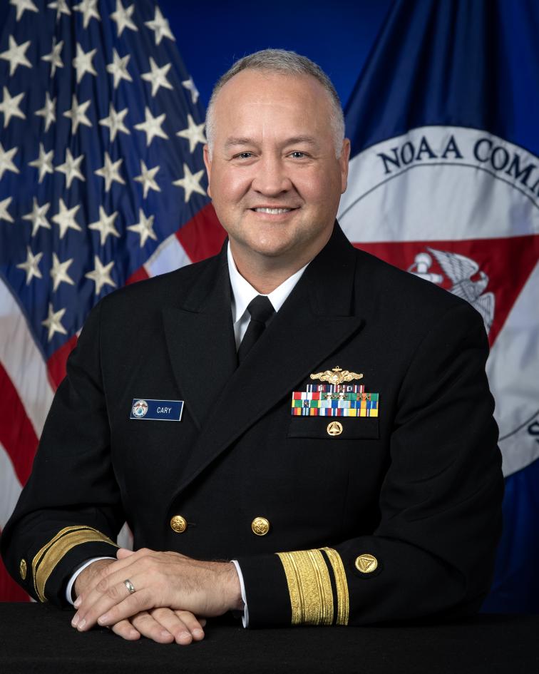 Rear Admiral Chad Cary in dress blue uniform with U.S. and NOAA Corps flags behind him