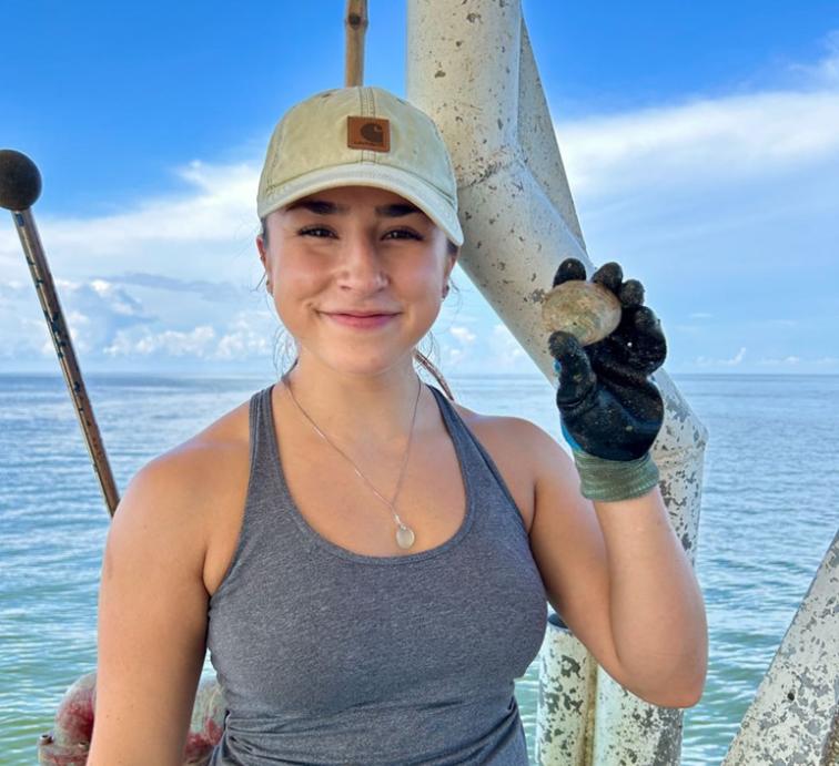 A woman smiles at the camera on a boat while holding a clam.