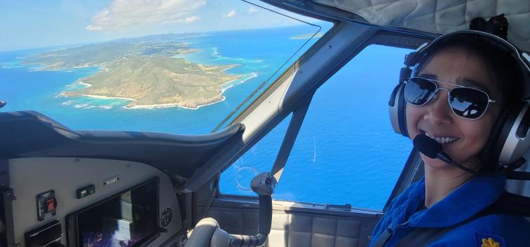 NOAA Corps pilot smiles in flight station of NOAA Twin Otter with view of St. Croix water and landscape in the background
