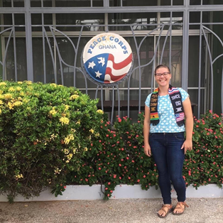A woman stands in front of a Peace Corps logo, smiling with a Peace Corps stole on.