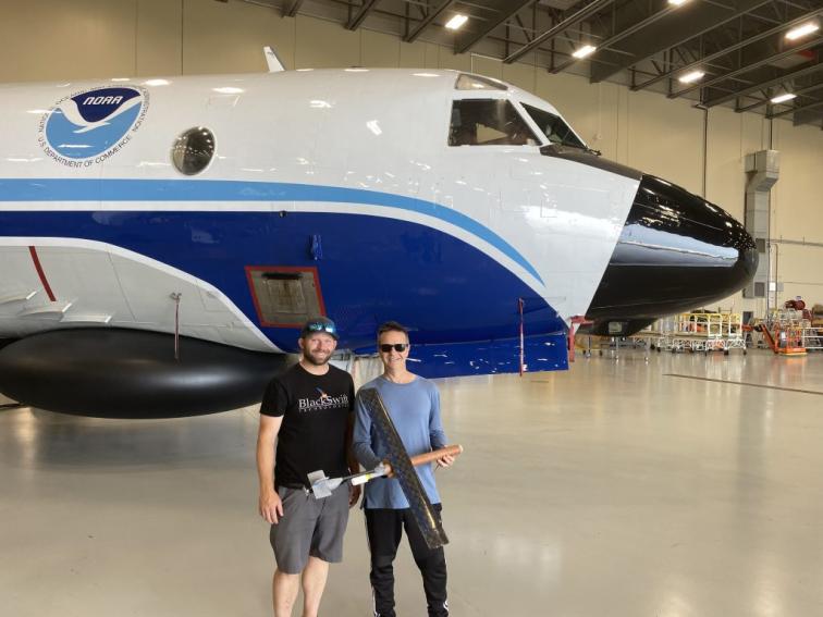 Two individuals stand in front of a NOAA Hurricane Hunter plane with a uncrewed aircraft in their hands.
