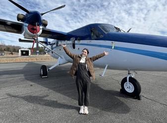 An OMAO Knauss Fellow stands in front of a NOAA Twin Otter plane, posing with a smile and outstretched arms.
