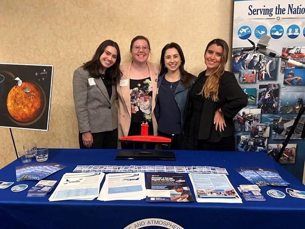 Four women in business casual attire stand in front of a table with NOAA outreach materials on it.