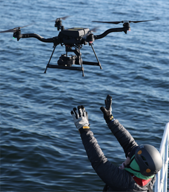 A person on the side of a ship extends their arms upwards, preparing to catch an uncrewed aircraft.